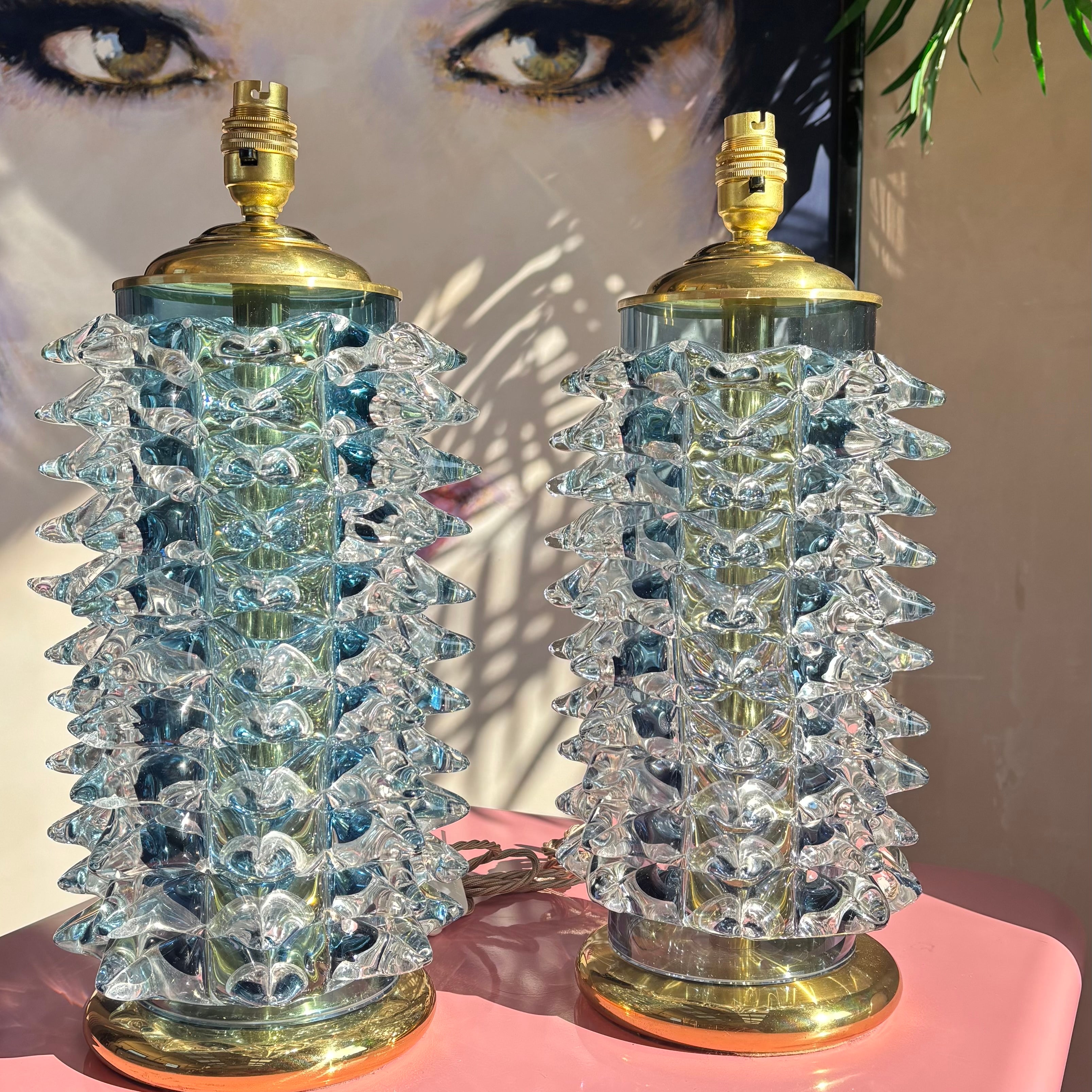Two Barovier and toso, murano lamps with crystal-spikes on a pink surface, with a large poster of a woman's face in the background. By Luigi Rocca 